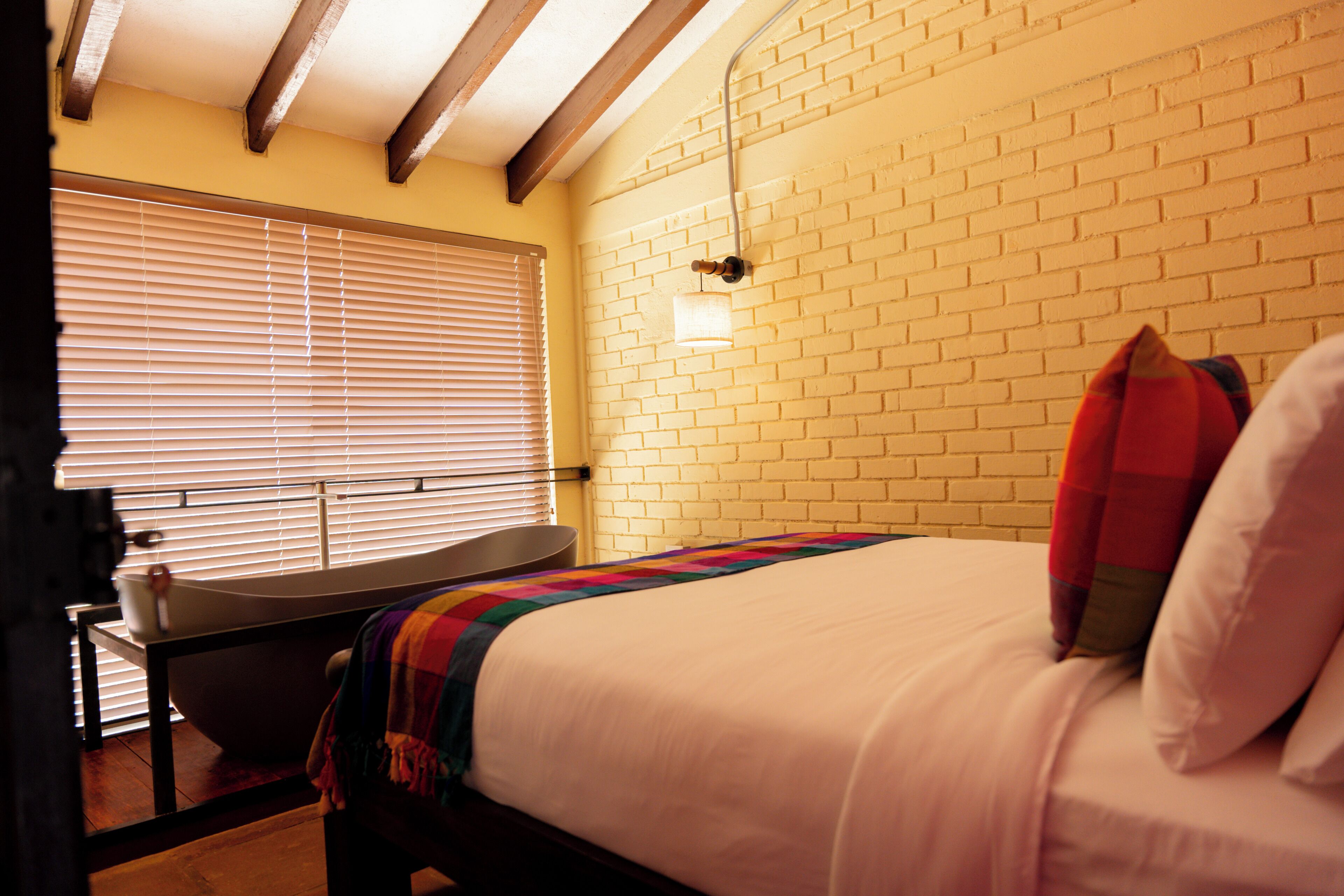 A bedroom with a bed, a bedside table, a window with blinds, a brick wall, and a light fixture.
