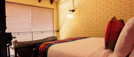 A bedroom with a bed, a bedside table, a window with blinds, a brick wall, and a light fixture.