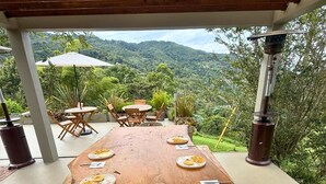 A long wooden table set for a meal with plates of food, utensils, and a view of a lush green landscape. - Hotel Campestre Rancho La Lechería (Cali)