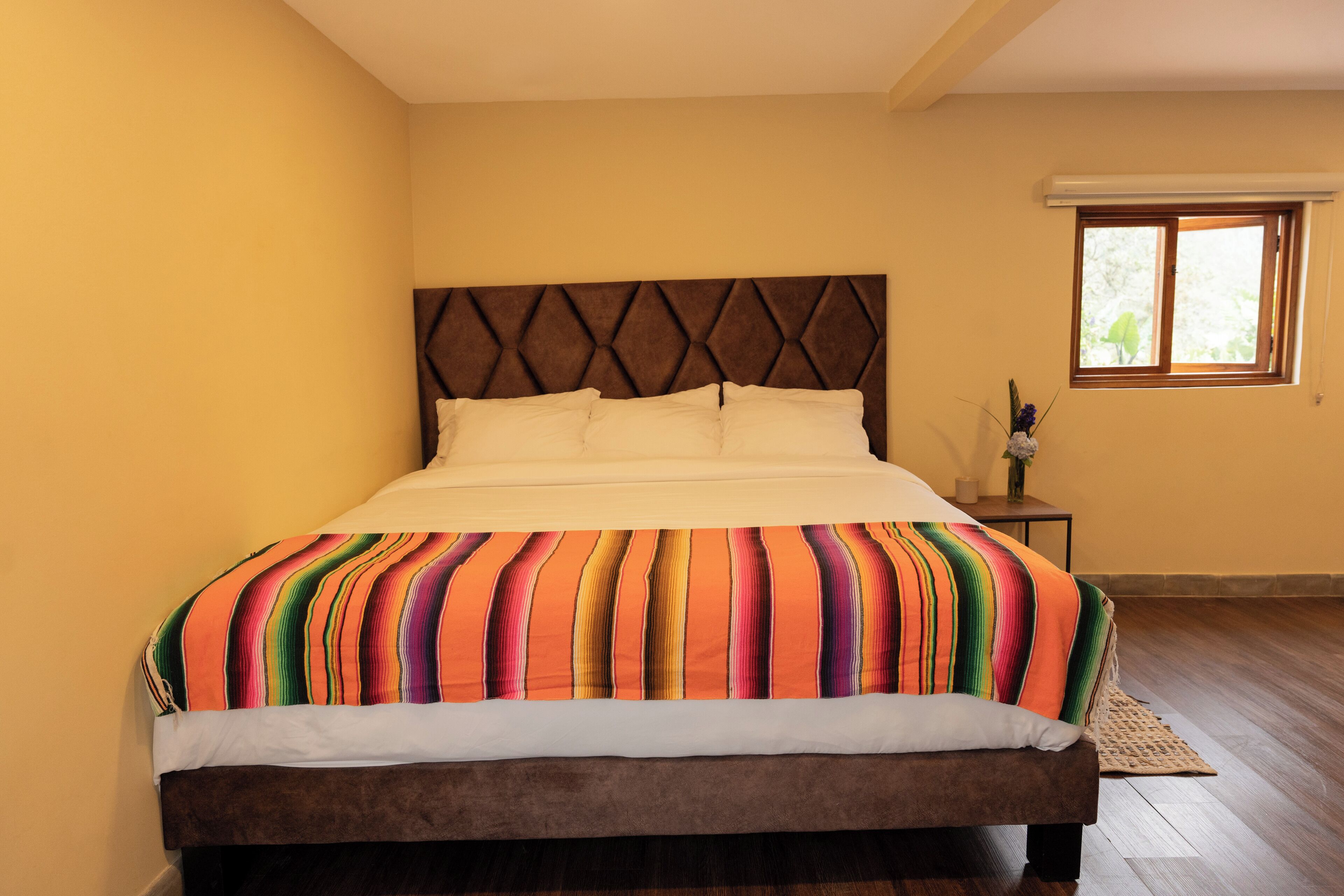 A bed with a colorful striped bedspread, a wooden headboard, and a window with a view of greenery.