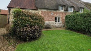 A traditional thatched-roof house with a brick exterior, a small window, and a wooden bench in the yard. - Bubbs Cottage (Dorchester)