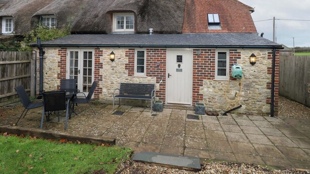 A stone cottage with a thatched roof, a white door, and a small garden with outdoor seating. - Bubbs Cottage (Dorchester)