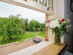 A view from a window with a table and chairs outside, a brick wall, and a vase with flowers on a wooden ledge. - Highfields Farm (Church Stretton)