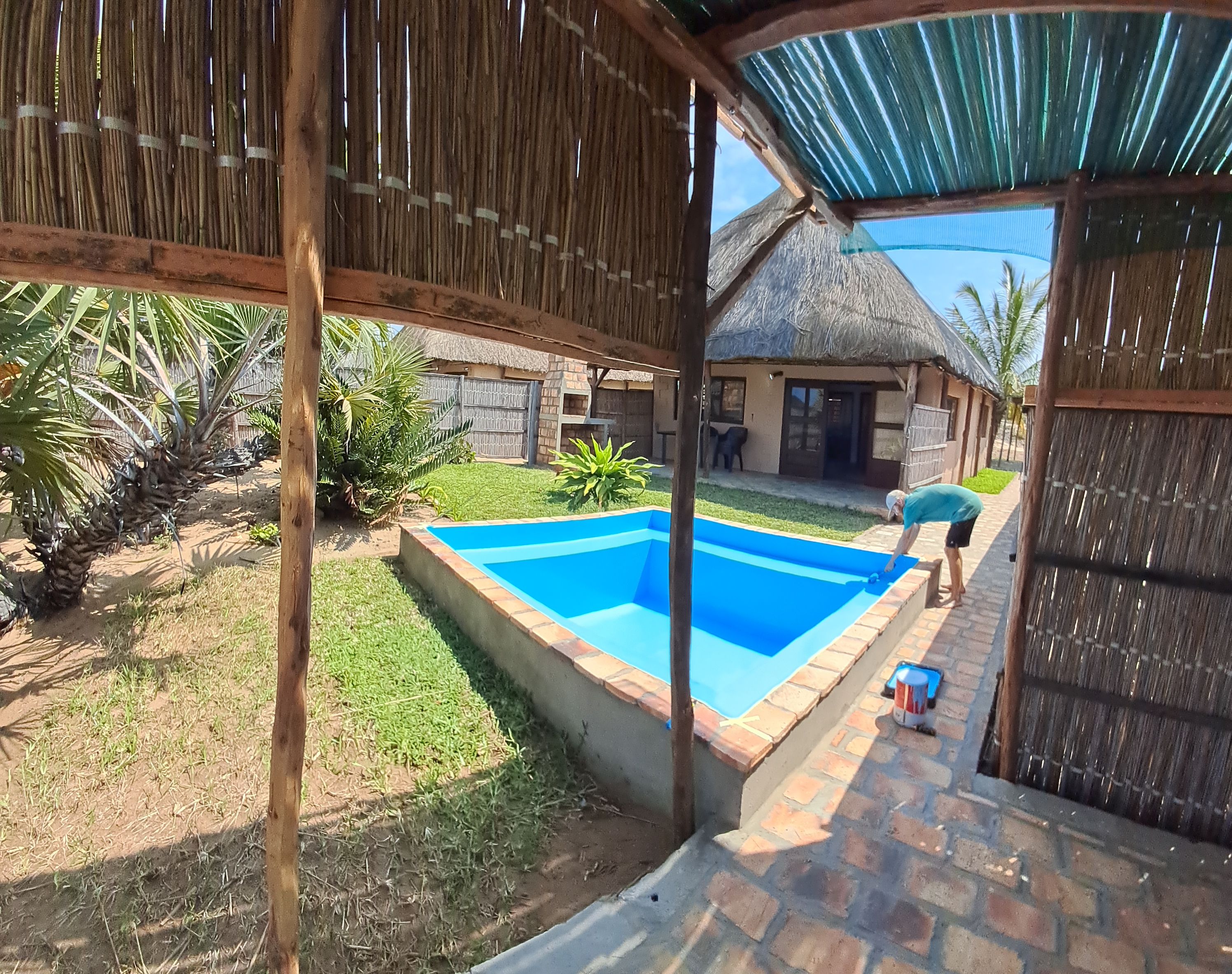 A backyard with a swimming pool, a thatched-roof structure, and a fenced area with vegetation.