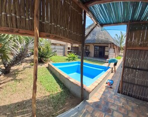 A backyard with a swimming pool, a thatched-roof structure, and a fenced area with vegetation.