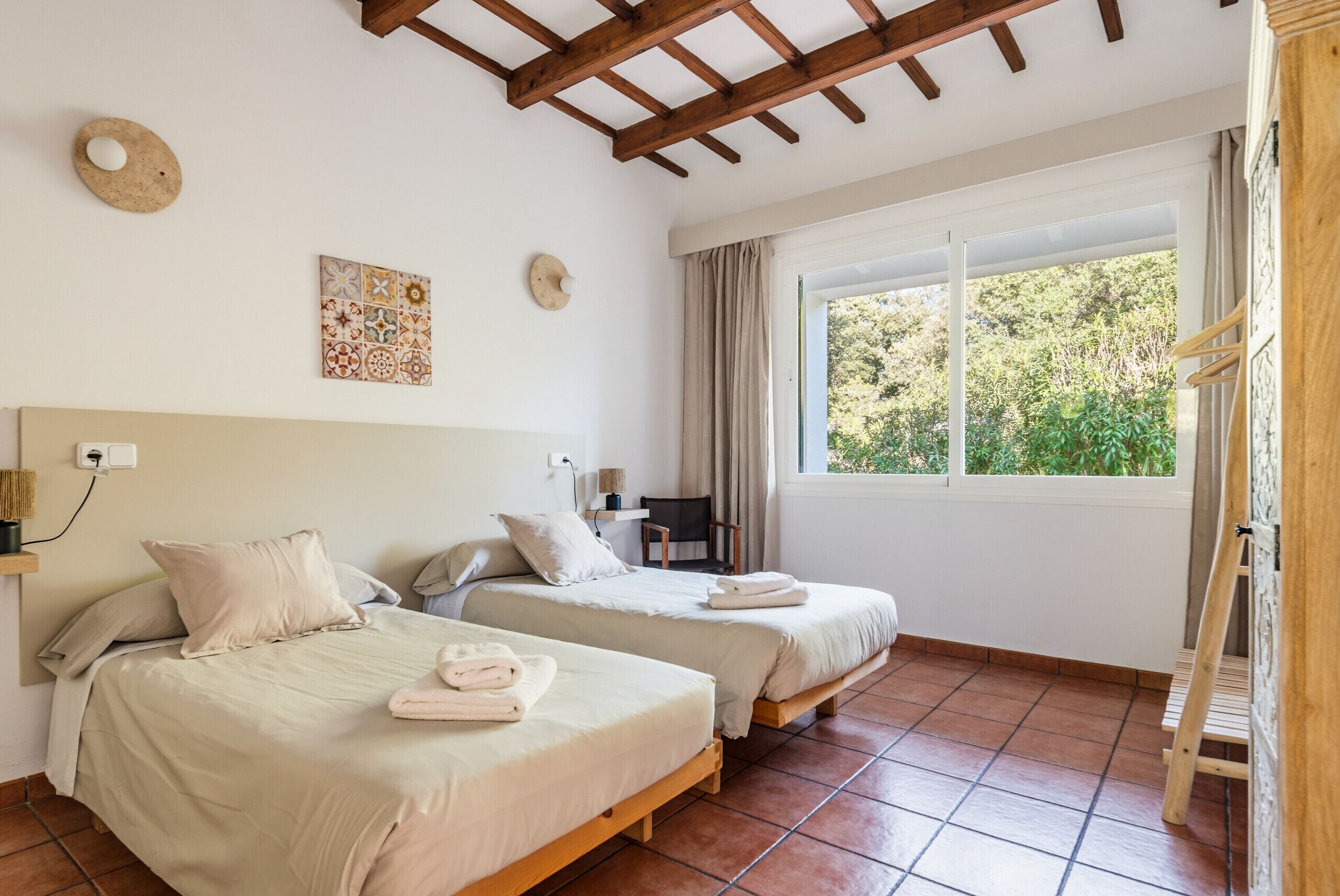 A room with two beds, a window with curtains, a wooden ceiling, and a wall-mounted light fixture.