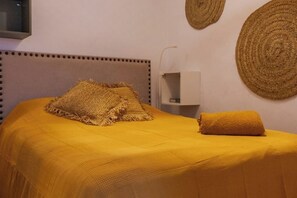 A bed with a yellow bedspread and two pillows, a bedside table with a lamp, and woven wall art. - Casa Bombón - Andalusian charm in the heart of the city (Medina-Sidonia)