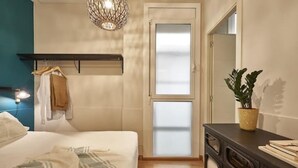 Une chambre à coucher comprenant un lit, une commode, un luminaire suspendu, une porte en verre dépoli et une plante en pot.