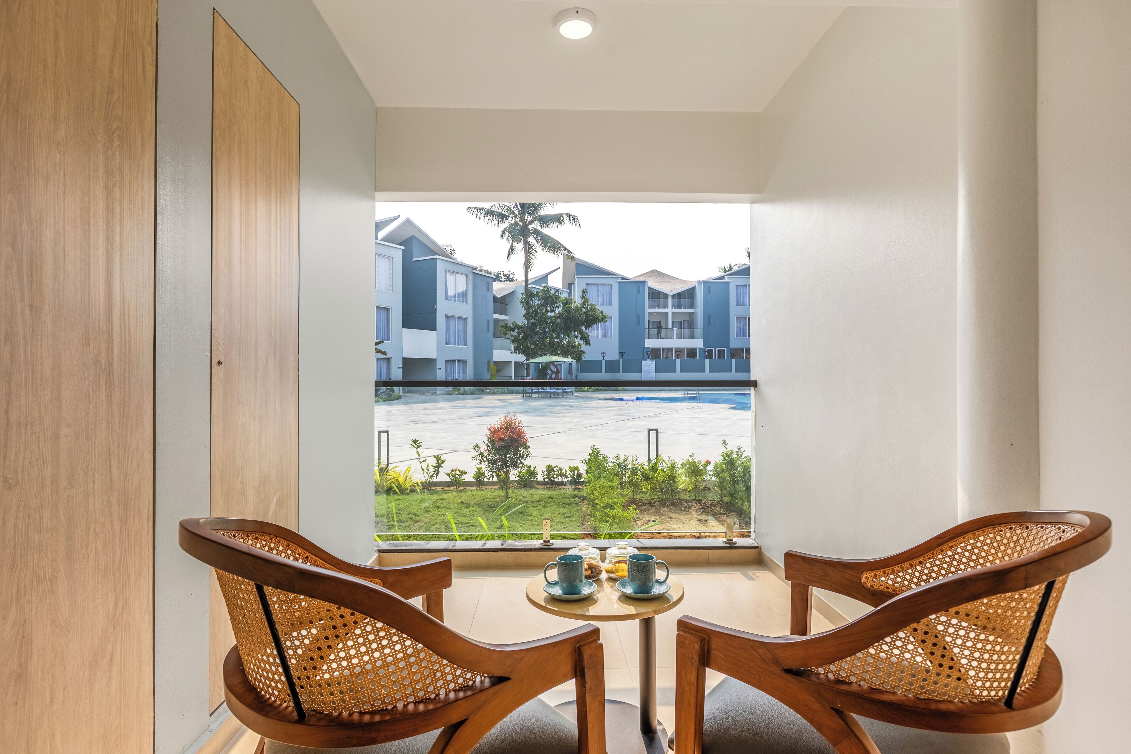 Varanda com duas cadeiras de vime e uma mesa pequena, com vista para uma piscina e edifícios do hotel.