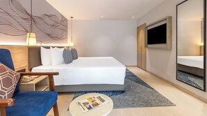 A modern hotel room with a large bed, a desk with a chair, a round coffee table, a large mirror, and a TV mounted on the wall. - Sterling Arka Suites Puri (Brahmagiri)