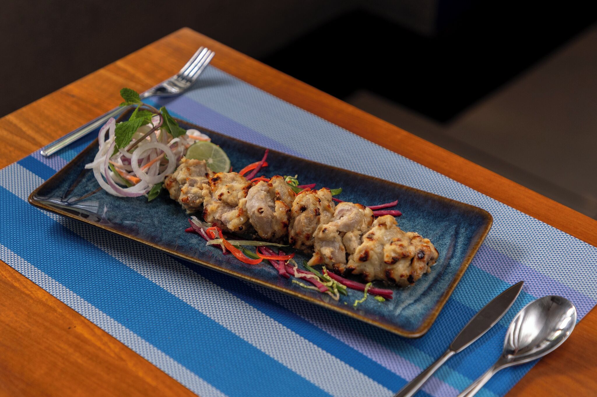 Un plato rectangular con brochetas de pollo a la parrilla, adornado con cebolla roja, lima y hierbas aromáticas, sobre un camino de mesa azul a rayas.
