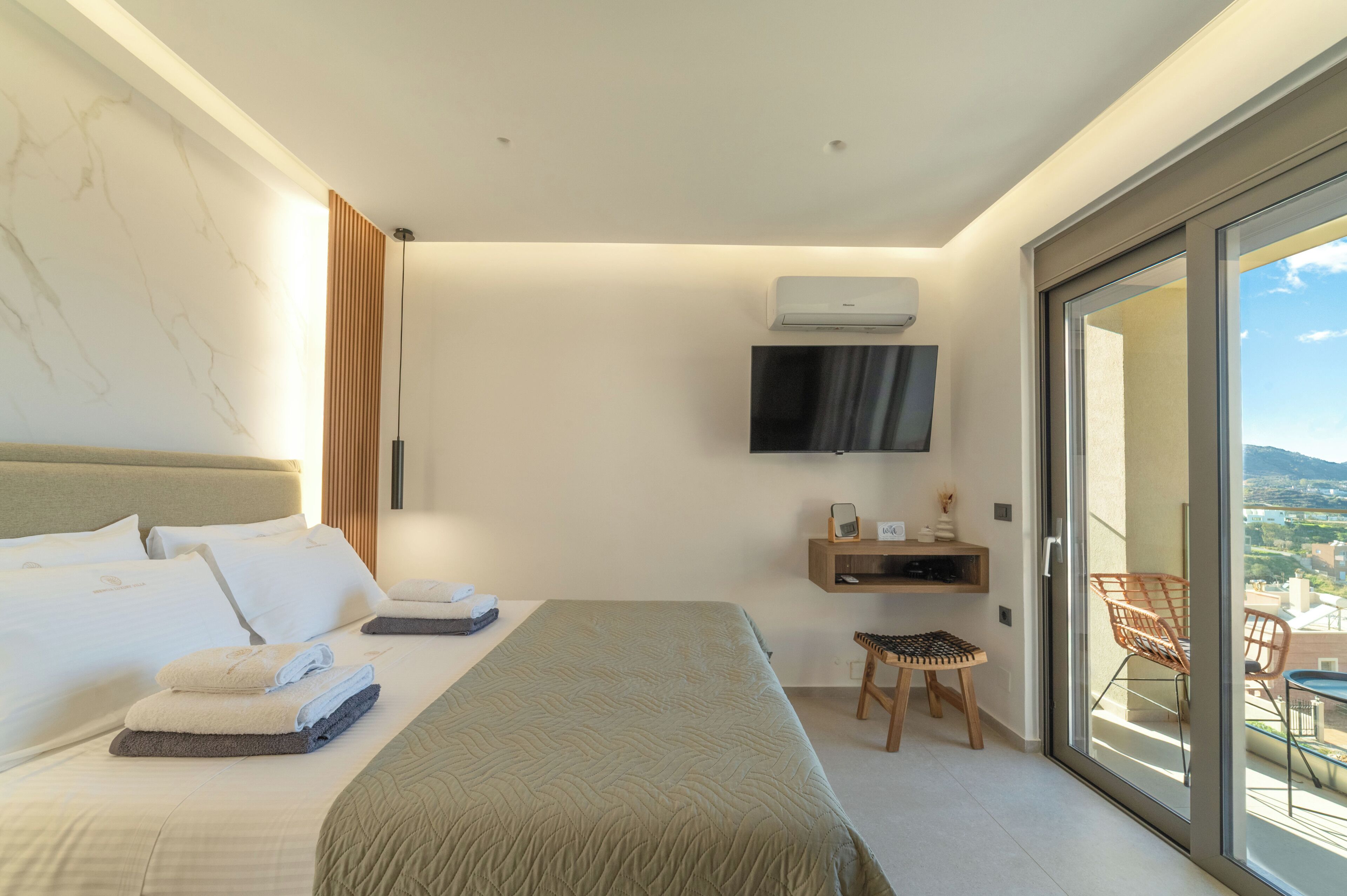 A modern hotel room with a large bed, a TV mounted on the wall, a small table with chairs, and a balcony with a view.