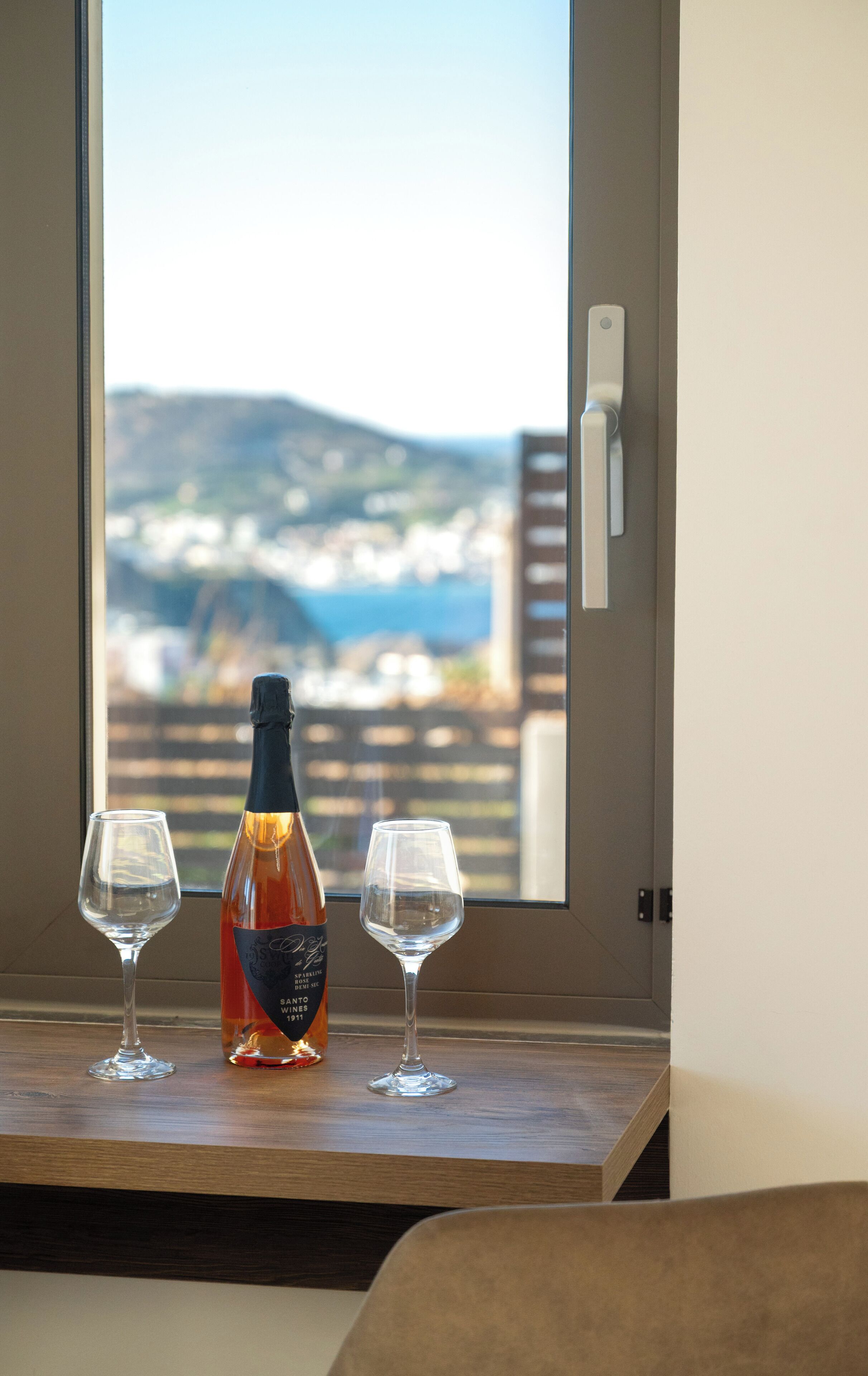 Uma garrafa de vinho e duas taças sobre uma mesa de madeira, perto de uma janela com vista para a paisagem costeira.