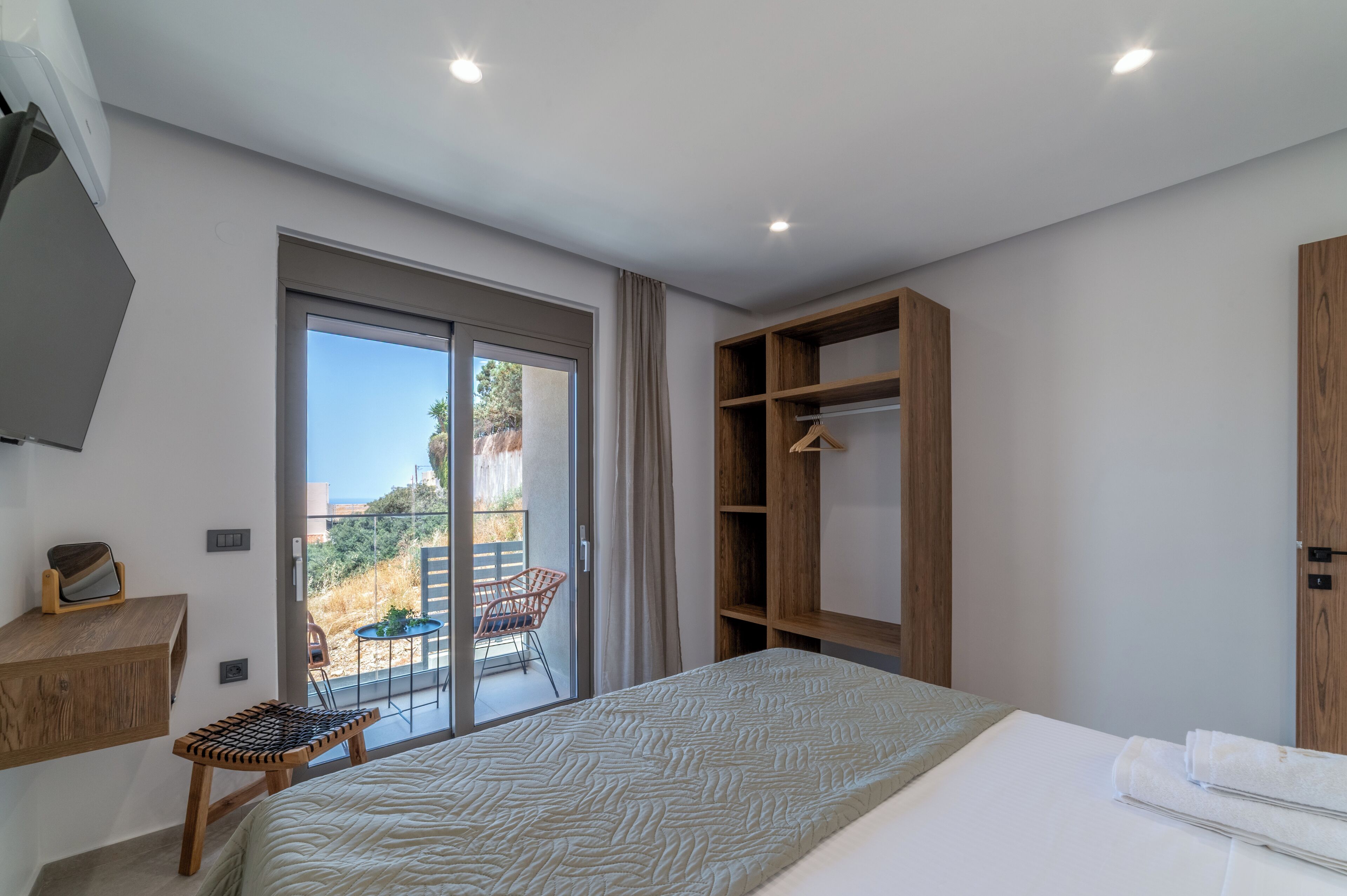 A bedroom with a bed, a wooden wardrobe, a desk with a chair, and a balcony with a table and chairs.