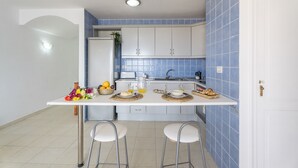 Cocina compacta con armarios blancos, salpicadero de azulejos azules, encimera blanca, dos taburetes de bar y un lavamanos pequeño.