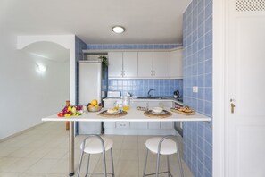 Een compacte keuken met witte kastjes, een blauwe betegelde spatwand, een wit aanrecht, twee barkrukken en een klein spoelbakje.