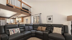 Un salon moderne avec un canapé d’angle en cuir noir, une cuisine en arrière-plan et une fenêtre offrant une vue agréable.