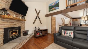 Un salon avec une cheminée, un canapé en cuir noir, un plancher en bois, un téléviseur fixé au mur et un tableau accroché au mur.