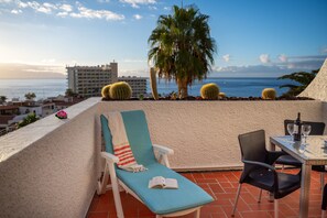 Un balcon avec un fauteuil de relaxation, une table avec des chaises et une vue sur l’océan et les bâtiments.