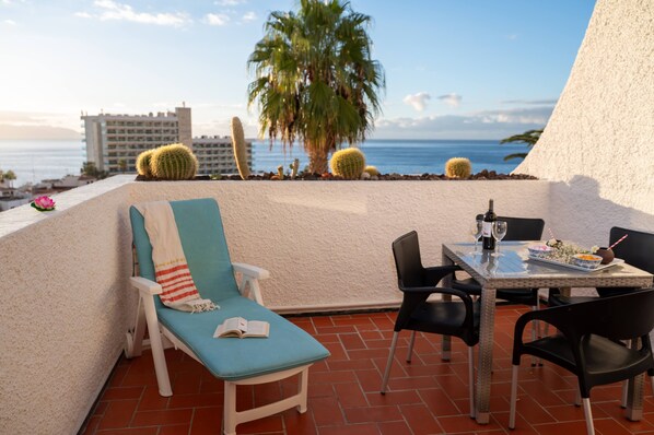 Une terrasse avec une table, des chaises, un fauteuil et une vue sur la mer et les palmiers.