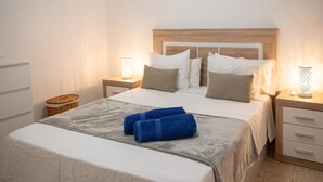 Una cama bien hecha con una colcha blanca y gris, dos toallas azules y un cabecero de madera.