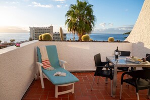 Un balcon avec vue sur la mer, une table avec une bouteille et des verres, et une chaise avec un livre.