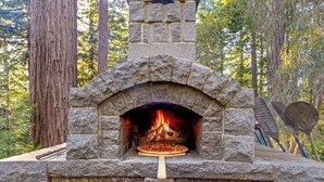 A stone-built pizza oven with a fire inside, surrounded by trees. - The Redstone House at Mendocino Stone Ranch (Anchor Bay)