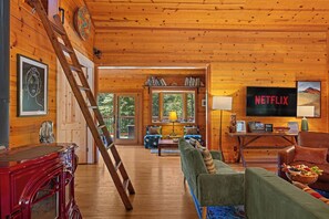 A wooden cabin interior with a living room, a staircase, a bookshelf, and a television.