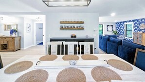 Eine moderne Küche mit einer zentralen Kochinsel, einem Essbereich mit einem weißen Tisch und gewebten Unterlagen, und ein blaues, gepolstertes Sofa im Hintergrund.