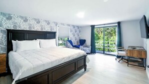 Ein Schlafzimmer mit einem großen Bett, einem Schreibtisch und einem Sessel. Ein Balkon bietet Blick auf Grünflächen.