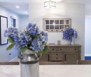 A dining area with a table, chairs, a vase with flowers, a wooden cabinet, and a wall-mounted light fixture. - Welcome to Plaisir d’Amour — “The Pleasure of Love”!  Sleeping 10! (Dennis)