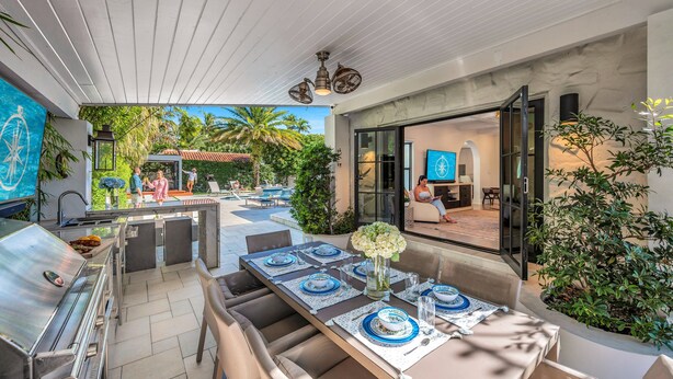 Ein Essbereich im Freien mit Grill, Tischen, die mit blau-weißen Tellern gedeckt sind, und Blick auf Pool und Haus im Hintergrund.