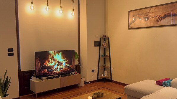 Een woonkamer met een flatscreentelevisie, een houten salontafel, een witte bank, een plant en een wandplank met decoratieve objecten.