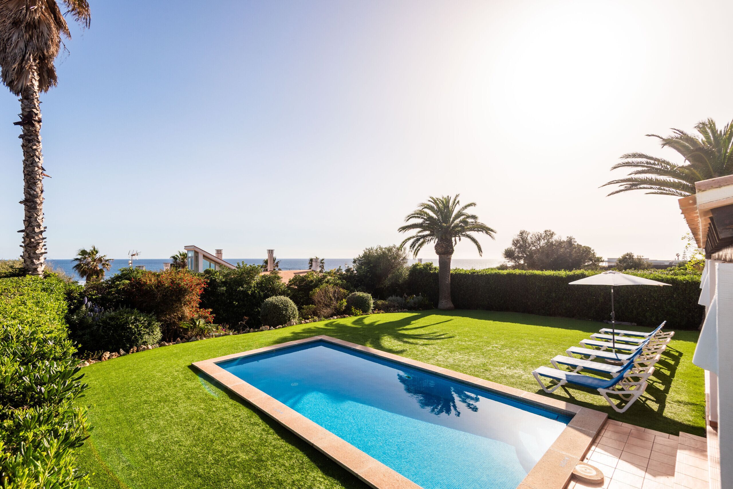 A swimming pool with a view of the sea, surrounded by well-maintained grass and palm trees.
