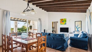 Ein Wohnzimmer mit Esstisch aus Holz, blauen Sofas, einem Fernseher und einer Decke mit sichtbaren Balken.