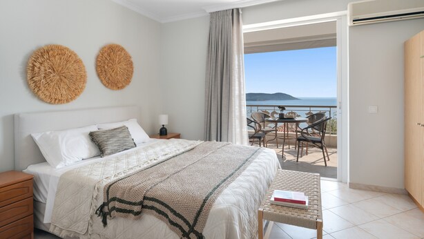 Ein Schlafzimmer mit Bett, Nachttischen, einer Nachttischlampe, Meerblick und einem Balkon mit Sitzgelegenheiten.