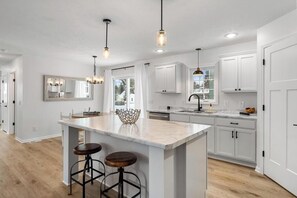 A modern kitchen with white cabinetry, a large island with stools, pendant lighting, and a view of the outdoors. - Uptown Retreat on Clark Street (Harbor Springs)