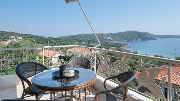Ein Balkon mit rundem Tisch und Stühlen, der auf eine Küstenlandschaft mit Hügeln und einem Gewässer blickt.