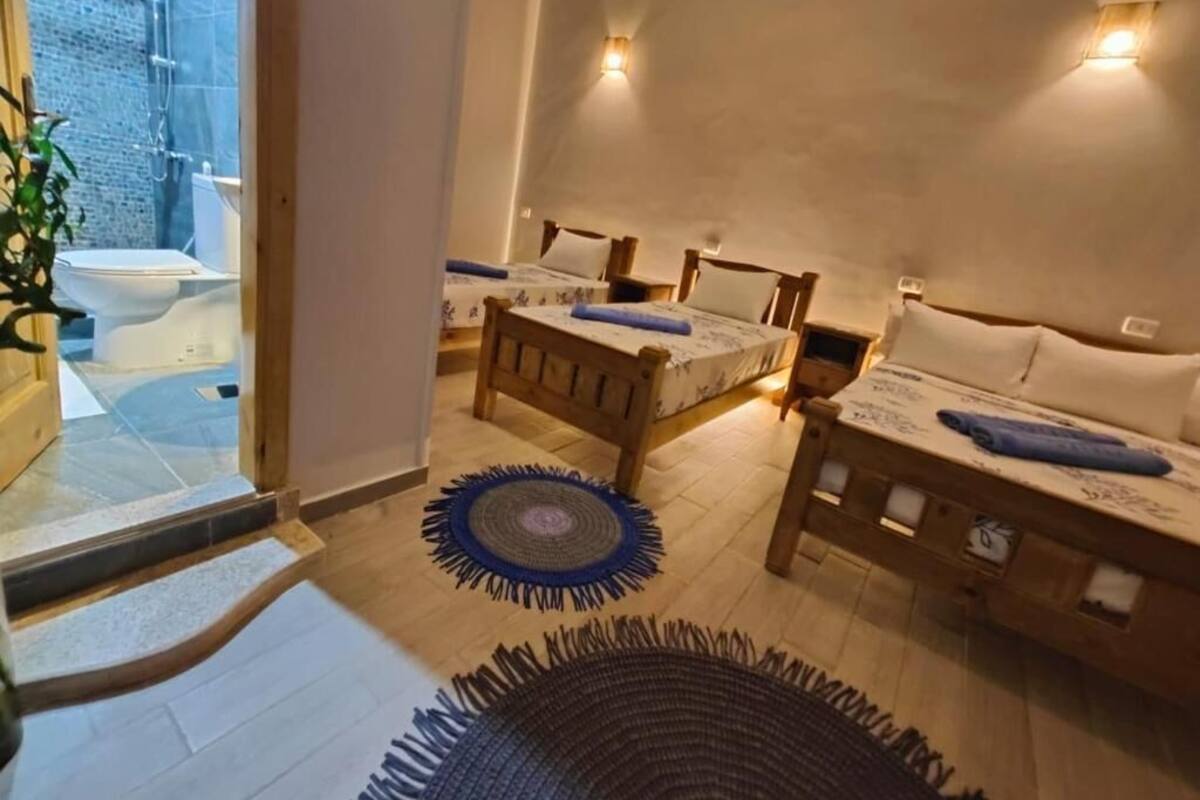 A cozy bedroom with two wooden beds, a small table, and a bench. There is a bathroom visible through an open door.