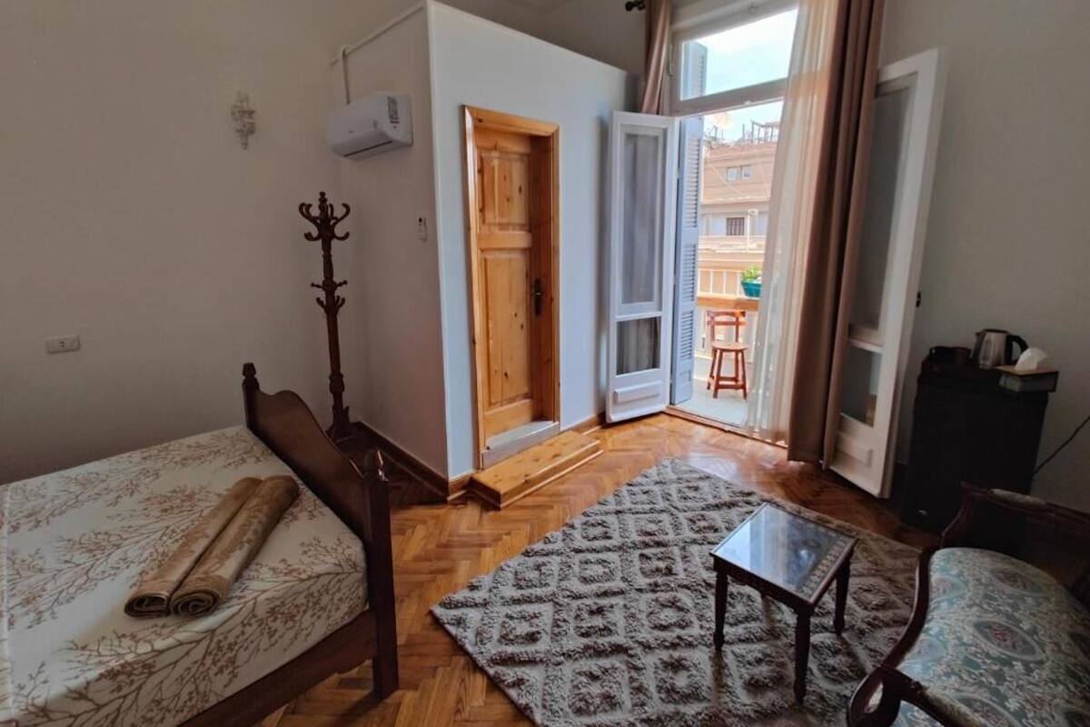 A small, furnished room with a bed, a wooden chair, a small table, and a patterned rug. There is a wooden door leading to an outdoor area.