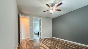 Una habitación amplia con piso de madera, ventilador de techo y puerta que da a un baño.