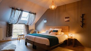 Un dormitorio con una cama grande, un cabecero de madera, una mesita de noche, un taburete pequeño y una ventana con cortinas.