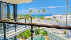 Varanda com guarda-corpo de vidro que dá para uma piscina, palmeiras e vista do oceano.