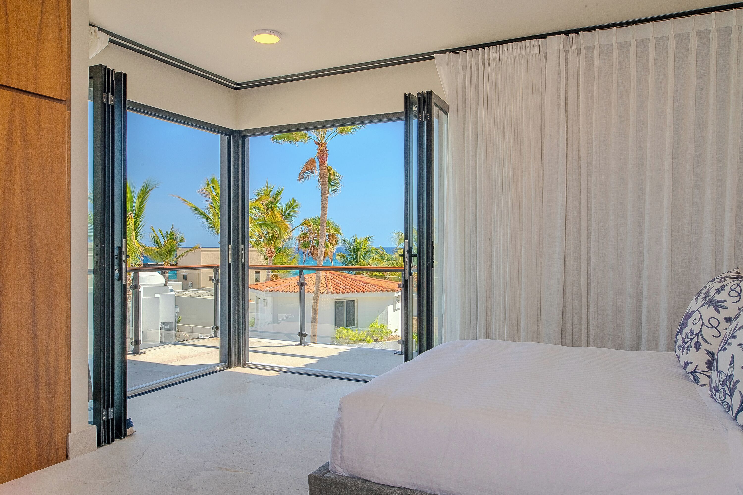A bedroom with a large bed, a view of palm trees and a building through open sliding doors, and a balcony with a railing.