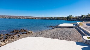 Un paseo marítimo con playa de guijarros, una zona de descanso de hormigón y vistas a un pueblo al otro lado del agua.