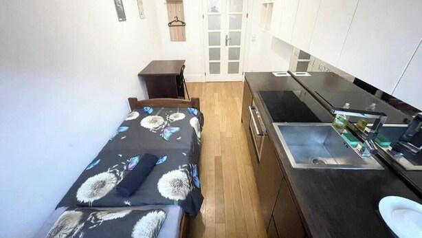 Una habitación compacta con cama, una mesita, una kitchenette con pileta y cocina, y una puerta que da a otra habitación.