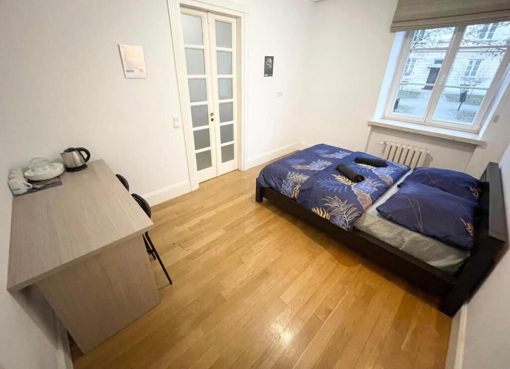 Un dormitorio con suelo de madera, una cama con colcha estampada, un escritorio con hervidor y una ventana con vistas a edificios.