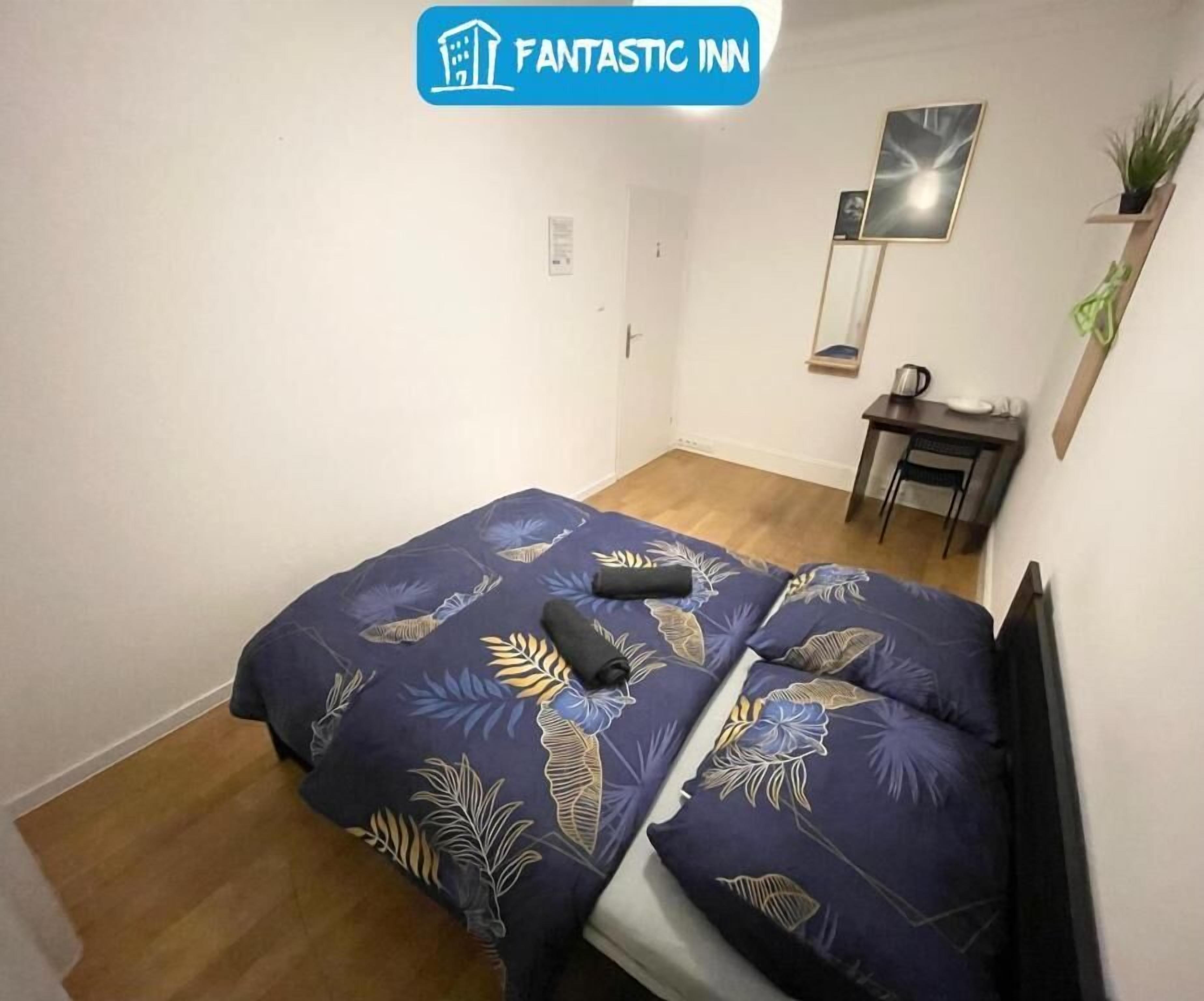 A small, single-bed room with a floral-patterned bedspread, a desk with a chair, and a small plant on the desk.