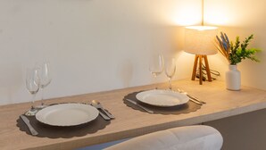 Comedor con dos mesas, cada una con un plato, cubiertos y dos copas de vino. Hay una lámpara con pantalla blanca y un jarrón con plantas secas. Dos cuadros enmarcados están colgados en la pared sobre las mesas.
