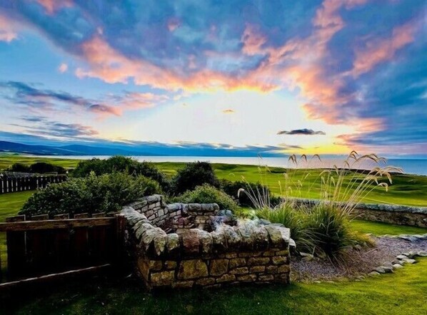 A stone wall with a rustic wooden fence, a grassy field, and a view of the sea at sunset.
